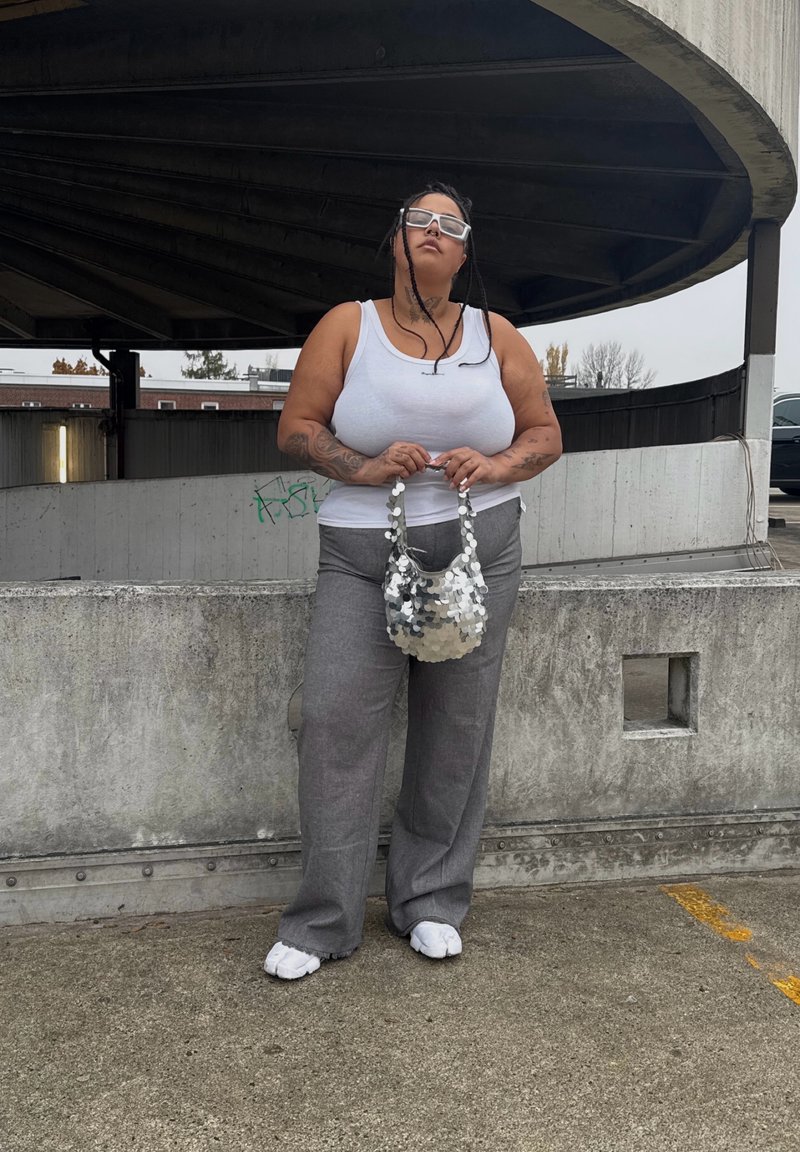 Gray wide-leg pants, white tank top, and a silver sequin handbag. She stands against a concrete backdrop with a textured surface.