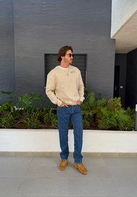 Beżowa bluza z nadrukowanym logo, niebieskie dżinsy, brązowe buty i okulary przeciwsłoneczne. Stoi przed ciemną ścianą i zielenią.