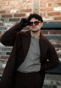 Homme aux cheveux bouclés ajuste ses lunettes de soleil, portant un manteau marron, un pull gris à torsades, des gants noirs, debout devant un mur en briques.