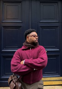 Man wearing glasses and a maroon sweater, standing with arms crossed and holding a brown leather bag, in front of a dark blue paneled door.