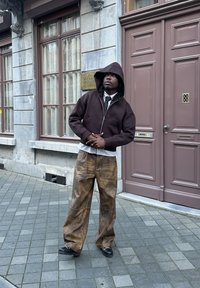 Brauner Reißverschluss-Hoodie mit Kapuze, über einem weißen Hemd und einer Krawatte getragen. Lockere, texturierte braune weit geschnittene Hosen und schwarze Schuhe. Urbane Umgebung.