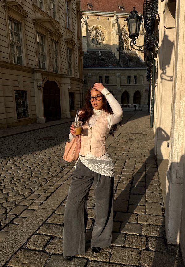 Νεαρή γυναίκα με γυαλιά κρατά κρύο καφέ και ροζ τσάντα, στέκεται σε πλακόστρωτο δρόμο κοντά σε ιστορικά κτίρια με ζεστό ηλιακό φως.