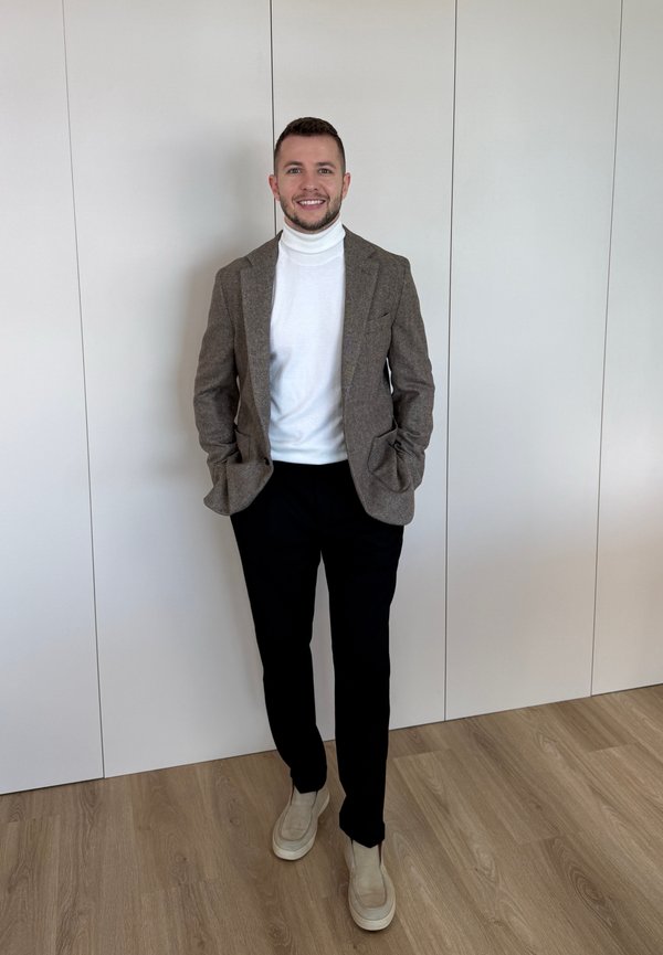Mann lächelt, trägt einen braunen Blazer, weißen Rollkragenpullover, schwarze Hosen und beige Schuhe, steht auf einem Holzboden vor weißen Wandpaneelen.