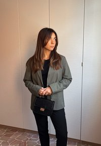 Sacou verde măsliniu peste un top negru, asortat cu pantaloni negri. Ține în mână o mică geantă neagră cu model de crocodil. Fundal deschis.