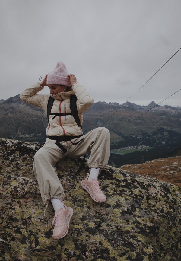 Chaqueta puffy ligera en crema con cremalleras de colores, pantalones cargo beige y zapatillas de deporte rosa pálido. La modelo está sentada sobre una superficie rocosa al aire libre.