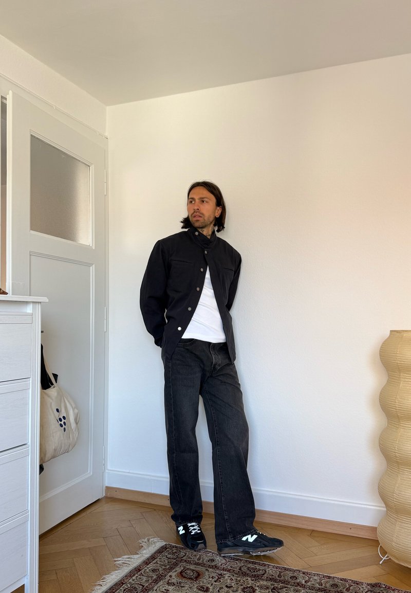 Homme aux cheveux mi-longs portant une veste noire, une chemise blanche, un jean noir et des baskets noires, debout à l'intérieur près d'une porte et d'une lampe.