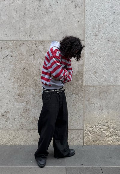 Persona con cabello rizado que lleva una sudadera con capucha a rayas rojas y grises, pantalones anchos negros y zapatos negros, de pie frente a una pared de piedra.