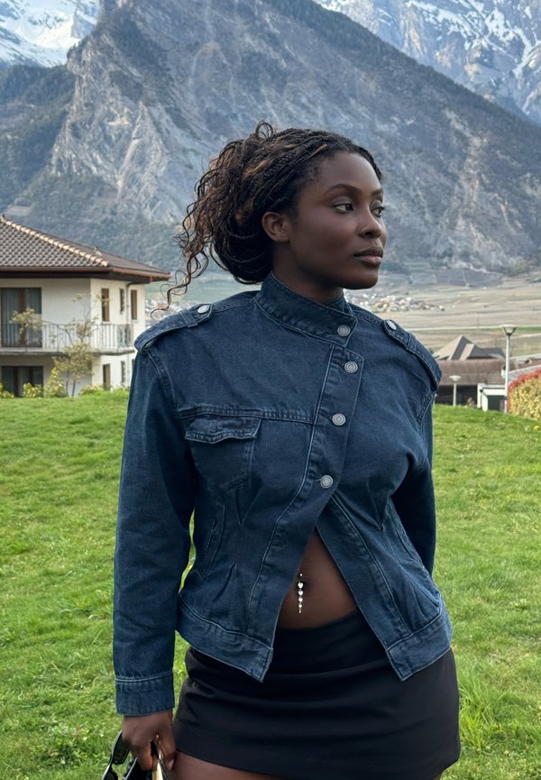 Femme aux cheveux tressés portant une veste en denim foncé et une jupe noire, debout dans une zone herbeuse avec des montagnes et des maisons en arrière-plan.