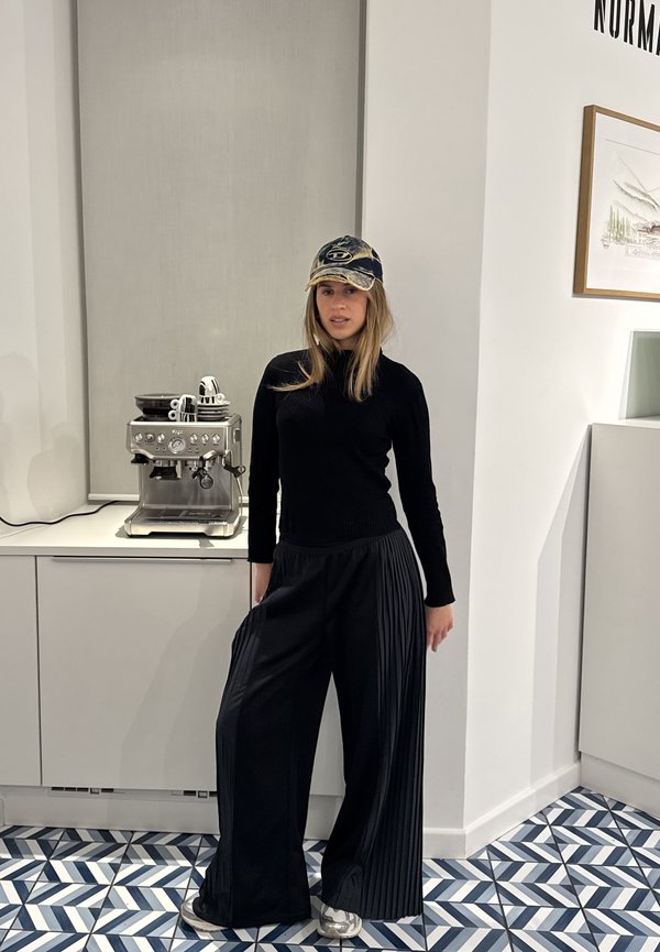 Tânără purtând un pulover negru, pantaloni largi plisati, șapcă de baseball și pantofi sport, stă într-o bucătărie modernă lângă o mașină de cafea.