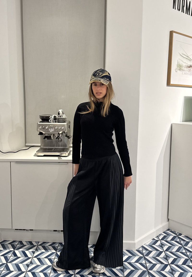 Mlada ženska, oblečena v črn pulover, široke hlače z naborki, kapo in superge, stoji v moderni kuhinji ob kavnem avtomatu.