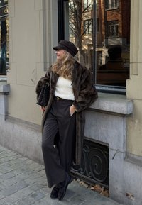 Γυναίκα με μαύρο καπέλο, καφέ γούνινο παλτό, λευκό πουλόβερ και μαύρο παντελόνι στηρίζεται σε γκρίζο πέτρινο κτίριο σε πλακόστρωτο δρόμο.