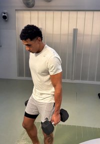 Homme soulevant des haltères dans une salle de sport, portant un t-shirt blanc et un short gris avec un tatouage sur le mollet gauche.