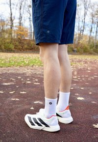 Baltas sporta kurpes ar melnām svītrām, kopā ar ceļgalu garuma tumši ziliem šortiem un baltām zeķēm. Apkārtne ir ar teksturētu, lapām klātu skrejceļu.