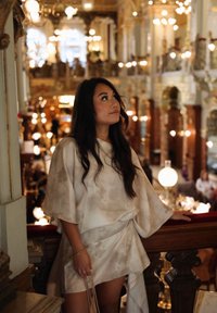 Une personne portant une tenue ample de couleur crème avec des motifs doux, se tenant près d'une balustrade en bois ornée dans un intérieur bien éclairé.