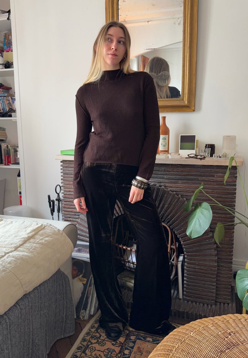 Pull marron foncé en tissu doux, associé à un pantalon large en velours noir. En arrière-plan, un miroir et des plantes d'intérieur.