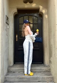 Pantaloni sportivi bianchi con strisce nere, una maglietta bianca, scarpe gialle, una borsa blu scuro e occhiali da sole, mentre tiene un gelato, vicino a una porta nera.