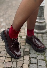 Zapatos burdeos de cordones con acabado brillante y suelas negras gruesas, llevados con calcetines rojos con volantes sobre una superficie de adoquines.