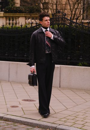 Chaqueta de cuero negra sobre una camisa de dress blanca y corbata estampada, combinada con pantalones negros y zapatos pulidos, sosteniendo un maletín negro.