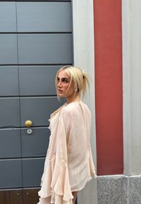Blusa ligera de un palo beige, con mangas de volantes fluidas y tela texturizada, combinada con una puerta gris y una pared roja de fondo.