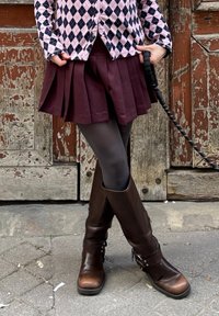 Γυναίκα στέκεται με σταυρωμένα πόδια, φορώντας μωβ πλισέ φούστα, πουλόβερ με διαμάντι σχέδιο, μαύρες καλσόν και καφέ μπότες μέχρι το γόνατο, κρατώντας το λουρί.