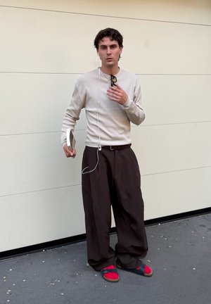 Joven con suéter beige, pantalones anchos marrones, calcetines rojos con sandalias negras, sosteniendo un libro y gafas de sol, de pie frente a una pared blanca.