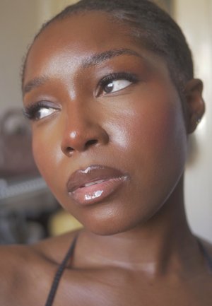 Close-up of a face with smooth, deep brown skin, glossy lips, and thick eyelashes. Light reflects softly, highlighting facial contours.