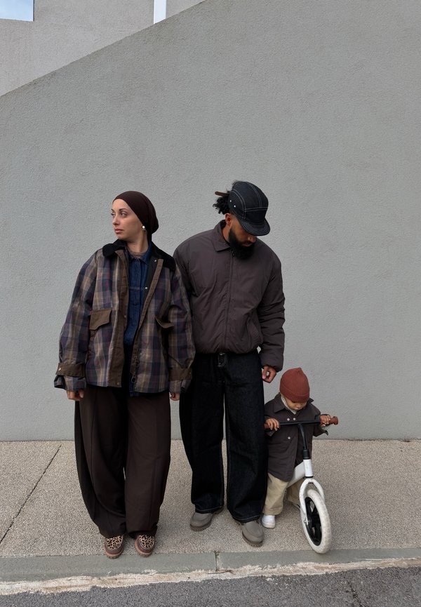 Dwoje dorosłych stojących obok małego dziecka trzymającego biały rowerek biegowy, wszyscy ubrani w codzienne jesienne ubrania na tle szarej ściany.
