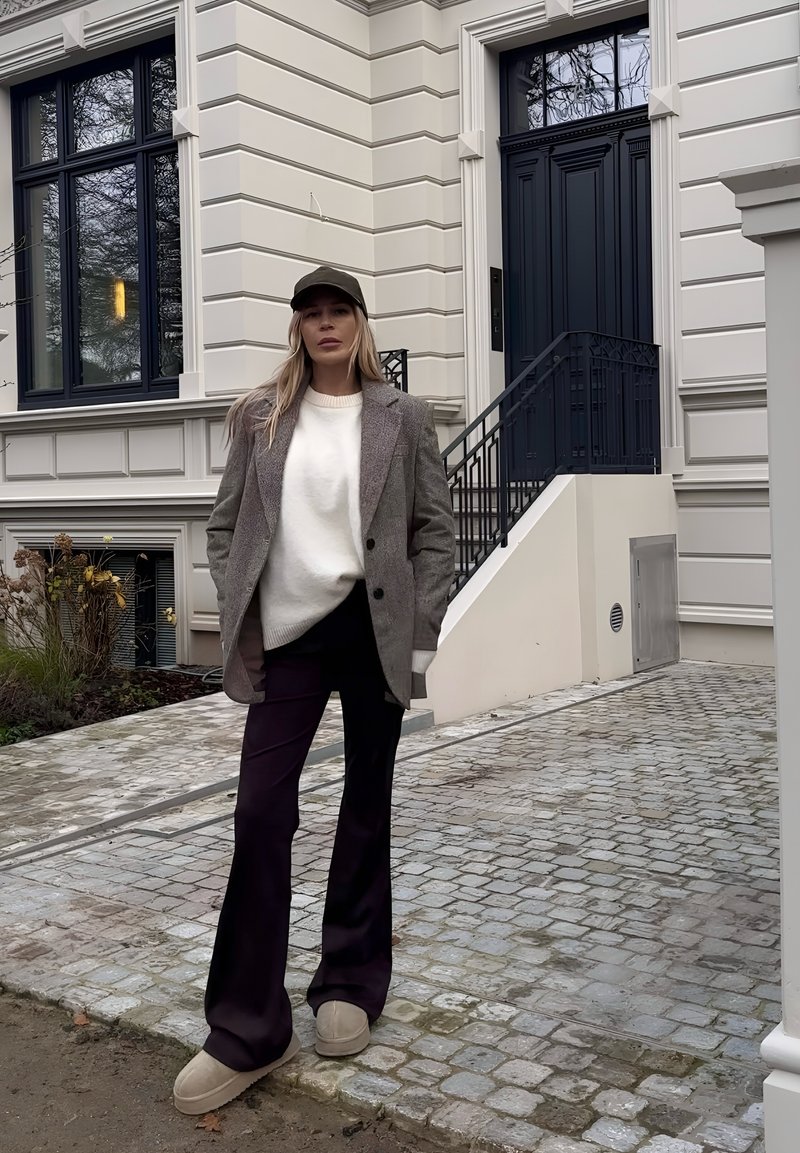 Femme debout sur un chemin pavé devant un bâtiment beige avec une porte noire, portant un blazer gris, un pull blanc, un pantalon noir et des chaussures beige.