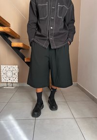 Μαύρο πουκάμισο με κουμπιά και λευκές ραφές, σε συνδυασμό με φαρδιά μαύρα σορτς. Μαύρες κάλτσες και παπούτσια ολοκληρώνουν το ντύσιμο. Ορατό γκρι πλακάκι στο πάτωμα.