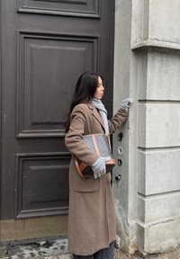 Casaco de lã castanho, cachecol cinzento e luvas cinzentas. Jeans azuis. Mala preta com acentos castanhos, junto a uma parede cinzenta texturizada e porta.