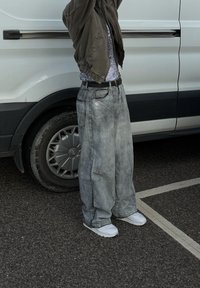 Laia, luhised hallist denimist teksad, mis on koos tumeda bomber-jakiga ja valgete tennisetega, seisab kõrvalt mikroauto.