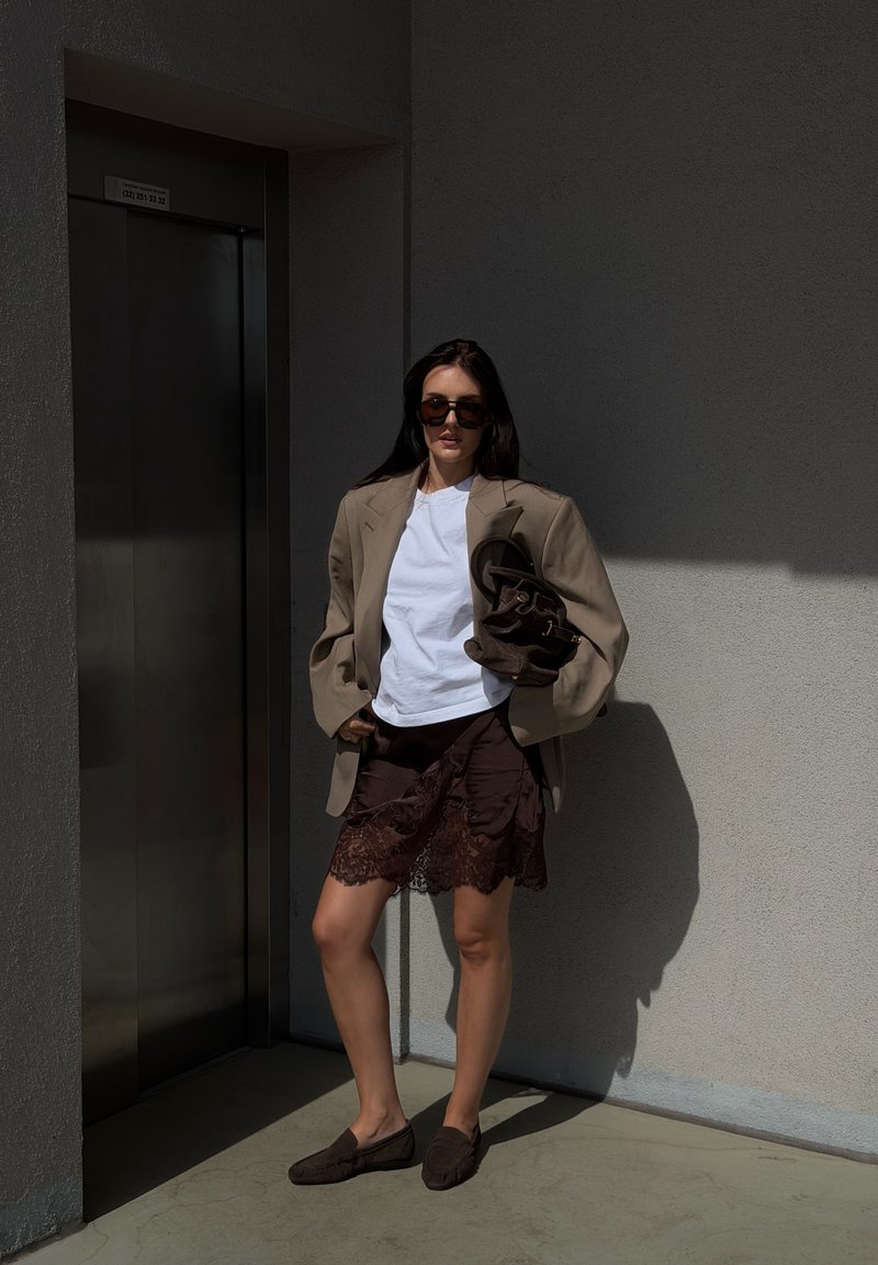 Mujer con camisa blanca, blazer beige, falda marrón de encaje, gafas de sol, sosteniendo bolso marrón, de pie a la luz del sol junto al ascensor y la pared gris.