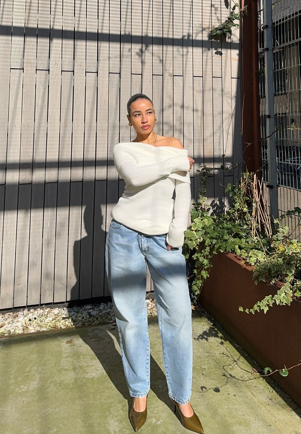 Weißer, gerippter Off-Shoulder-Pullover, hellblaue lässige Jeans und dunkelgrüne, spitze Absätze, vor einem schrägen Holzhintergrund mit grünem Bewuchs.