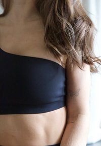 Czarny top crop na jedno ramię o gładkiej fakturze, z dopasowanym fasonem podkreślającym talię. Na ramieniu widoczny jest mały tatuaż motyla.