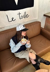 Femme portant une chemise rayée, un gilet noir et une casquette, assise sur un canapé marron, tenant une tasse de latte avec une mousse en forme de cœur, près d'une tenture murale intitulée « Le toile ».