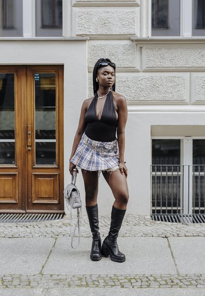 Cuerpo de halter negro combinado con una falda corta de cuadros, botas de plataforma negras hasta la rodilla y una bolsa gris. Superficies texturizadas y colores contrastantes.
