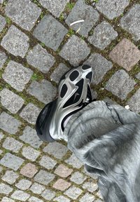 Fekete és ezüst sneaker, hálós felsőrésszel, jellegzetes ovális kivágásokkal és ívelt talppal, egy köves térkövezett felületen elhelyezve.