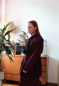 A red-haired woman wearing a maroon blazer and skirt stands beside a wooden cabinet adorned with potted plants and a lamp in a softly illuminated room.
