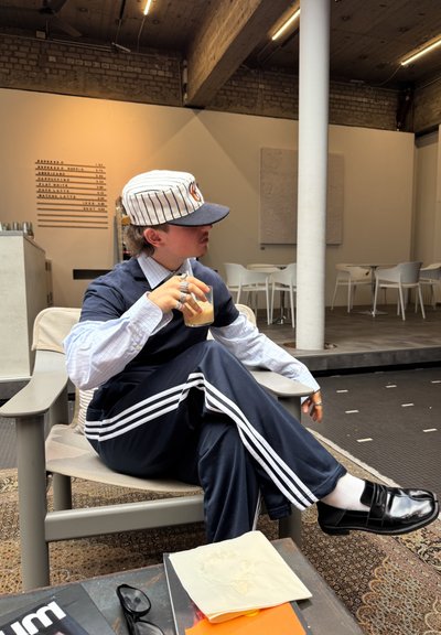 Persona vestida con un chándal azul marino con rayas blancas, sosteniendo una bebida, sentada en una silla. Lleva una gorra de béisbol a rayas y zapatos negros brillantes.