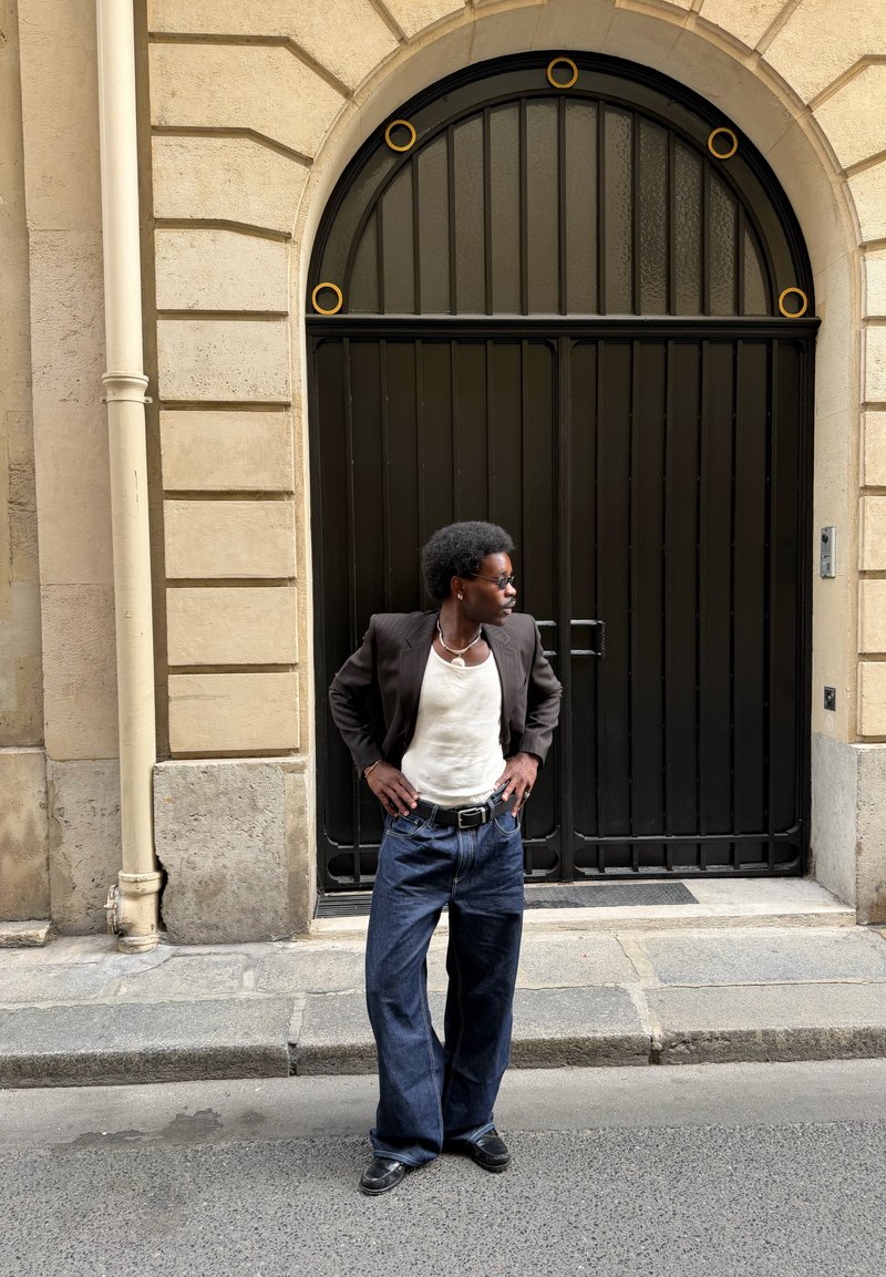 Hombre con peinado afro, gafas de sol, blazer oscuro, camisa blanca y vaqueros anchos, está de pie en la calle frente a una gran puerta negra arqueada.