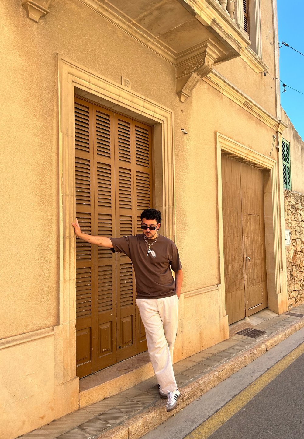 Braunes kurzärmeliges Hemd, beigefarbene Hose und Sneakers; steht neben einer Holzlamellentür vor einer sandfarbenen Steinmauer.