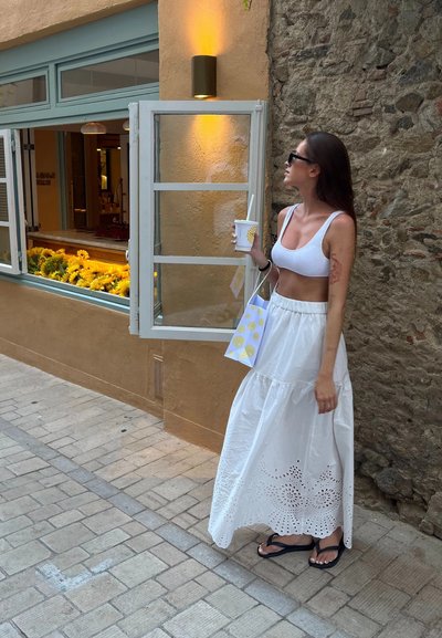 Una mujer con un top corto blanco y una larga falda blanca con detalles de ojal, sosteniendo una bebida y una bolsa de compras con estampado. Fondo de pared de ladrillo.