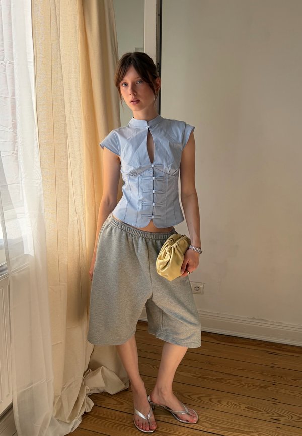 Jonge vrouw staat binnen, draagt een lichtblauw mouwloos topje, losse grijze shorts, zilveren sandalen, en houdt een beige clutch vast.