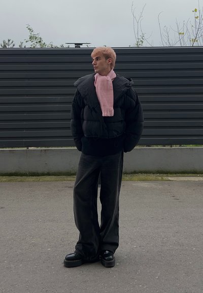 Chaqueta negra acolchada con cuello alto, bufanda rosa, vaqueros oscuros y zapatos negros robustos. De pie frente a un fondo de madera oscura.