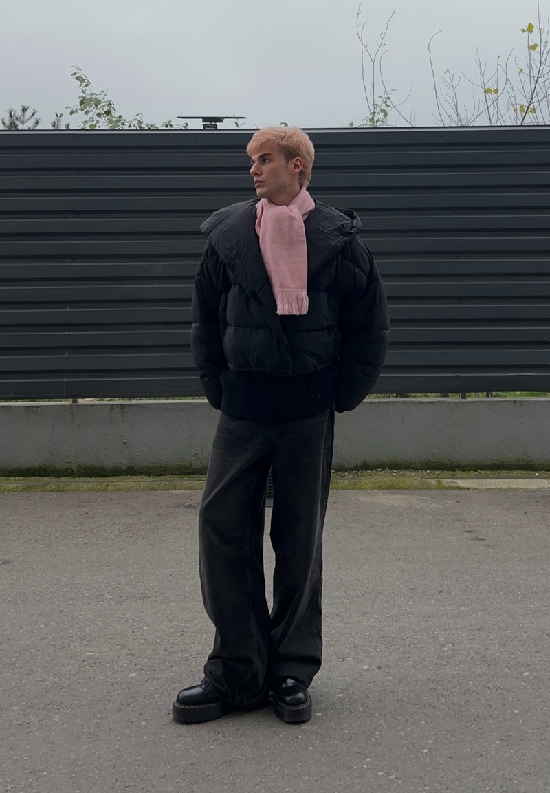 Doudoune noire à col montant, écharpe rose, jean plus foncé et chaussures noires massives. Debout devant un fond en bois sombre.