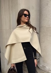 Beige wollen poncho met een asymmetrisch ontwerp, elegant vallend over een zwart shirt met lange mouwen, gecombineerd met zwarte nauwsluitende broek.