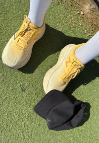 Oseba v rumenih tekaških copatih in belih nogavicah stoji na travi ob črni kapici znamke Under Armour.