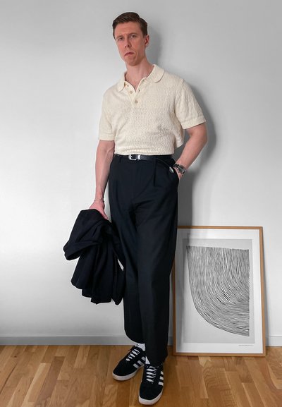 Hombre con polo crema texturizado, pantalones negros y zapatillas sostiene chaqueta negra, de pie junto a una obra de arte abstracta en blanco y negro con marco en el suelo.