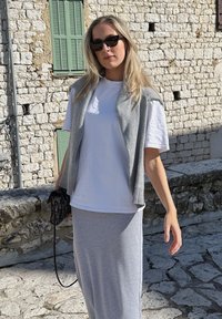 Grijze mouwloze trui over een wit t-shirt, gecombineerd met een lange grijze rok. Gecombineerd met een kleine zwarte tas. Lichte achtergrond van een stenen muur.