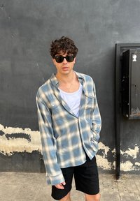 Camicia in flanella a scacchi blu e beige con maniche lunghe, indossata aperta sopra una canotta bianca, abbinata a pantaloni corti neri e occhiali da sole scuri.